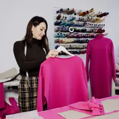 Krawcowa w pracowni z mnóstwem nici. Na wieszaku wisi bluza w intensywnym kolorze magenta, jaki to kolor! Obok na manekinie sukienka w tym samym odcieniu.