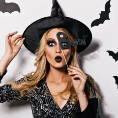 Kobieta w kapeluszu czarownicy i błyszczącej sukience, z wyrazistym makijażem, trzyma tabliczkę "Boo!".