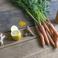 Na drewnianym stole leżą marchewki, imbir, cytryna, butelka soku i rozsypane **plamy z kurkumy**.