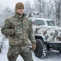 Mężczyzna w zimowej kurtce moro i czapce stoi na tle pojazdu wojskowego w śnieżnym krajobrazie. Idealna kurtka na zimę.