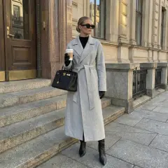 Eleganckie stylizacje z płaszczem: szary, długi płaszcz z paskiem, czarna torebka i wysokie kozaki. Kobieta z kawą na mieście.