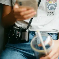 Osoba w jeansach z wysokim stanem (mid waist) trzyma telefon i napój. Na szyi wisi aparat.