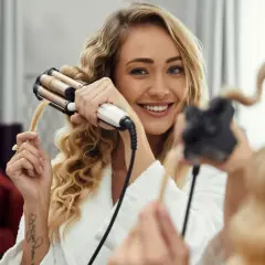 Uśmiechnięta blondynka pokazuje, jak zrobić fale na włosach za pomocą karbownicy. Jej włosy są pięknie pofalowane.