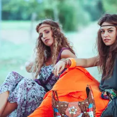 Dwie dziewczyny w stylu moda lat 70. siedzą na trawie, otoczone naturą. Jedna w kwiecistej sukience, druga w luźnej tunice.