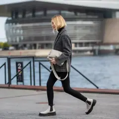 Kobieta w czarnych spodniach i marynarce, z jasną torebką, prezentuje stylowe, białe botki na grubej podeszwie. Idealne buty do czarnych spodni damskich.