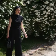 Kobieta w czarnym, eleganckim stroju, jak prac czarne pranie, stoi na tle kwitnących krzewów.