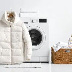 Biała kurtka puchowa wisi obok pralki, gotowa na pranie. W koszyku obok znajduje się detergent do prania.