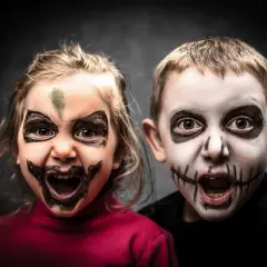 Dwoje dzieci z przerażającymi makijażami na Halloween. Dziewczynka ma zieloną plamę na czole i czarne wzory wokół oczu. Chłopiec ma białą twarz z czarnymi szwami na ustach. To łatwy makijaż na Halloween dla dzieci.