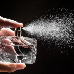 Dłoń trzyma szklaną buteleczkę, z której rozpyla się mgiełka. To woda perfumowana, lekka jak perfumy.