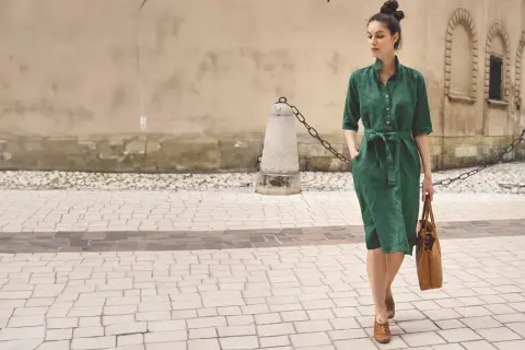 Elegancka kobieta w zielonej lnianej sukience prezentuje letnie stylizacje do biura, idąc ulicą z brązową torbą.
