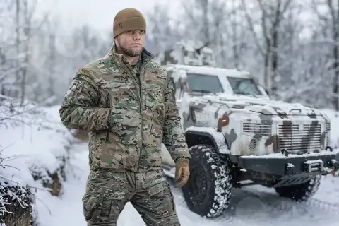 Mężczyzna w zimowej kurtce moro i czapce stoi na tle pojazdu wojskowego w śnieżnym krajobrazie. Idealna kurtka na zimę.