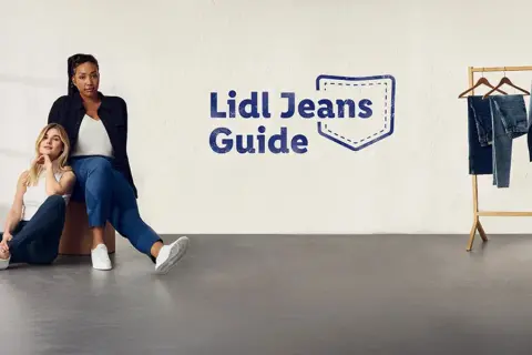 Dwie kobiety w jeansach siedzą na podłodze. Obok wieszak z jeansami. Rozmiar 27 jaki to rozmiar? Sprawdź w Lidl Jeans Guide.