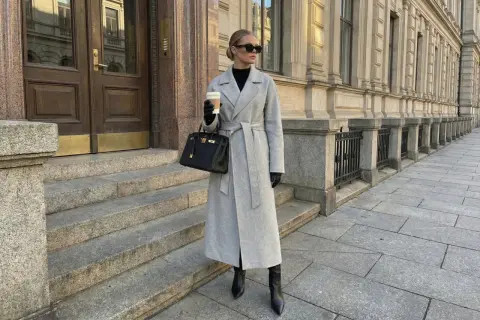 Eleganckie stylizacje z płaszczem: szary, długi płaszcz z paskiem, czarna torebka i wysokie kozaki. Kobieta z kawą na mieście.