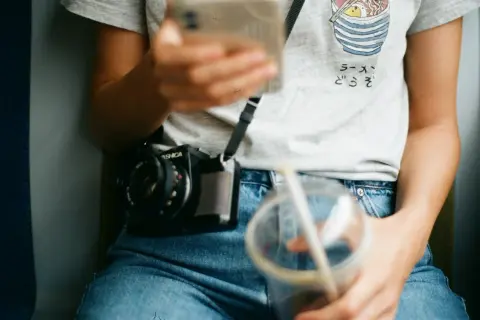 Osoba w jeansach z wysokim stanem (mid waist) trzyma telefon i napój. Na szyi wisi aparat.