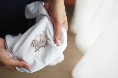 Dłonie trzymają białą tkaninę z widocznymi, **starymi tłustymi plamami na ubraniach**.