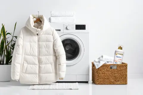 Biała kurtka puchowa wisi obok pralki, gotowa na pranie. W koszyku obok znajduje się detergent do prania.