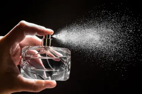 Dłoń trzyma szklaną buteleczkę, z której rozpyla się mgiełka. To woda perfumowana, lekka jak perfumy.