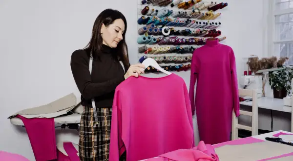 Krawcowa w pracowni z mnóstwem nici. Na wieszaku wisi bluza w intensywnym kolorze magenta, jaki to kolor! Obok na manekinie sukienka w tym samym odcieniu.