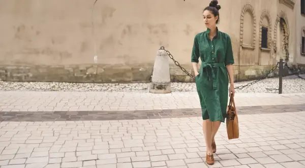 Elegancka kobieta w zielonej lnianej sukience prezentuje letnie stylizacje do biura, idąc ulicą z brązową torbą.