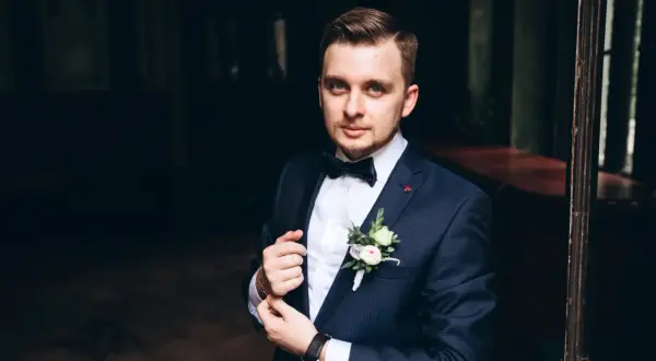 Elegancki mężczyzna w granatowym garniturze, z butonierką i muszką. Idealny przykład, jak dobrać garnitur na specjalną okazję.