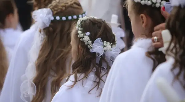 Dziewczynki w białych strojach z pięknymi fryzurami komunijnymi z wiankiem z kwiatów i wstążek.