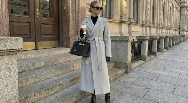Eleganckie stylizacje z płaszczem: szary, długi płaszcz z paskiem, czarna torebka i wysokie kozaki. Kobieta z kawą na mieście.