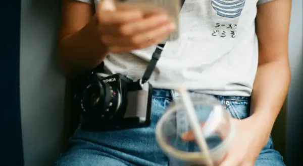 Osoba w jeansach z wysokim stanem (mid waist) trzyma telefon i napój. Na szyi wisi aparat.