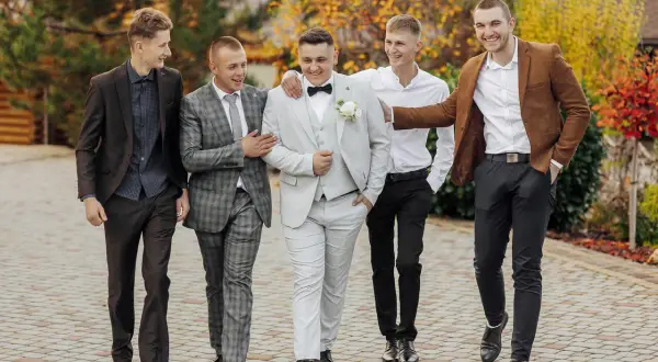 Pięciu mężczyzn w eleganckich stylizacjach na wesele, idących razem i rozmawiających.