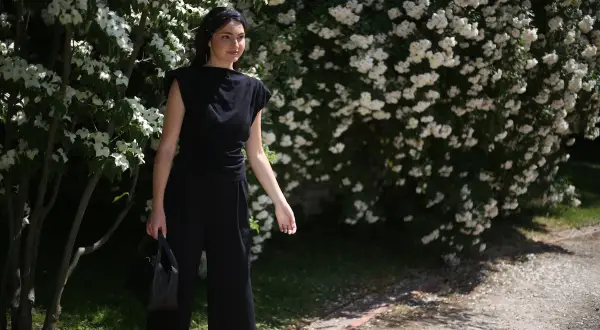 Kobieta w czarnym, eleganckim stroju, jak prac czarne pranie, stoi na tle kwitnących krzewów.