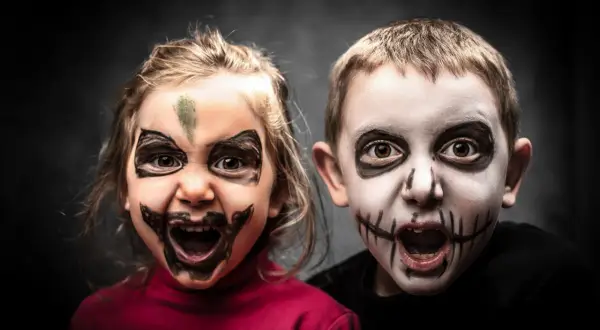 Dwoje dzieci z przerażającymi makijażami na Halloween. Dziewczynka ma zieloną plamę na czole i czarne wzory wokół oczu. Chłopiec ma białą twarz z czarnymi szwami na ustach. To łatwy makijaż na Halloween dla dzieci.
