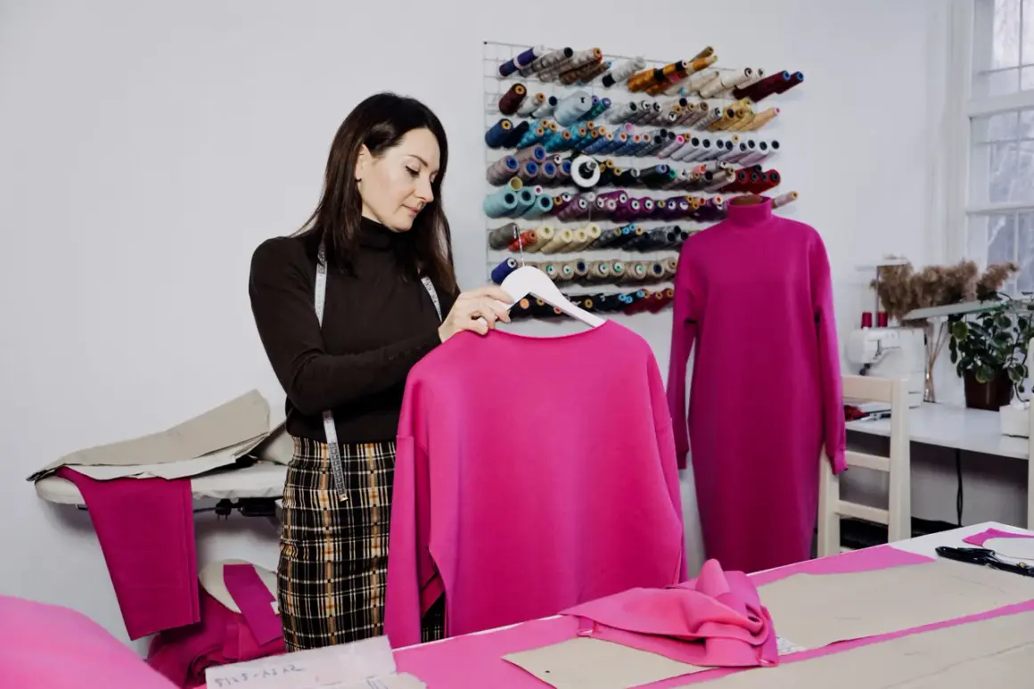 Krawcowa w pracowni z mnóstwem nici. Na wieszaku wisi bluza w intensywnym kolorze magenta, jaki to kolor! Obok na manekinie sukienka w tym samym odcieniu.