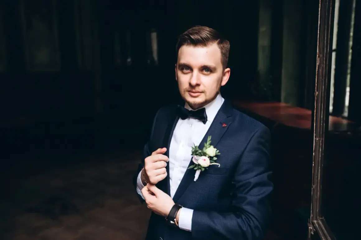 Elegancki mężczyzna w granatowym garniturze, z butonierką i muszką. Idealny przykład, jak dobrać garnitur na specjalną okazję.