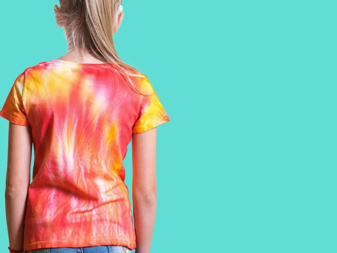 Dziewczynka w koszulce tie-dye w odcieniach czerwieni i żółci. To znaczy, że wzór powstał przez wiązanie i farbowanie materiału.