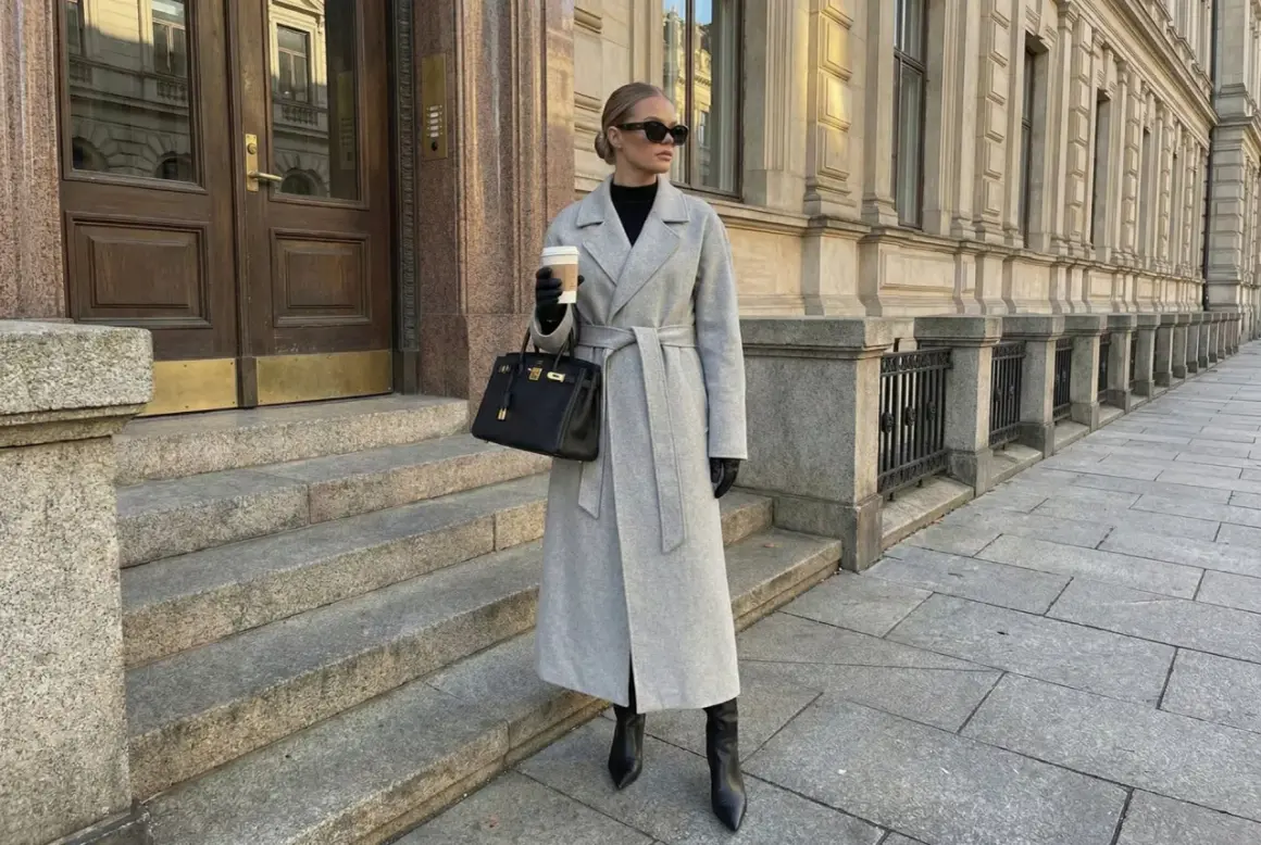 Eleganckie stylizacje z płaszczem: szary, długi płaszcz z paskiem, czarna torebka i wysokie kozaki. Kobieta z kawą na mieście.