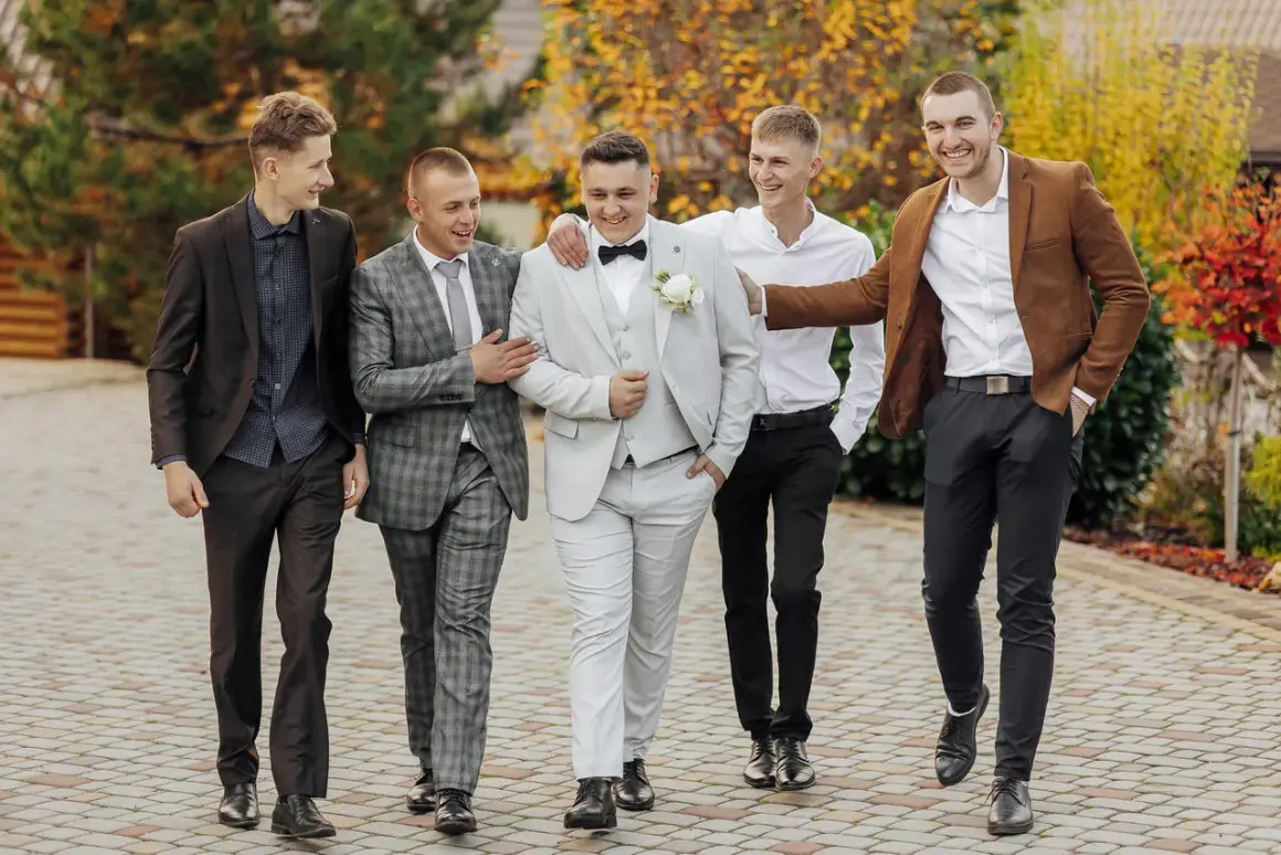 Pięciu mężczyzn w eleganckich stylizacjach na wesele, idących razem i rozmawiających.