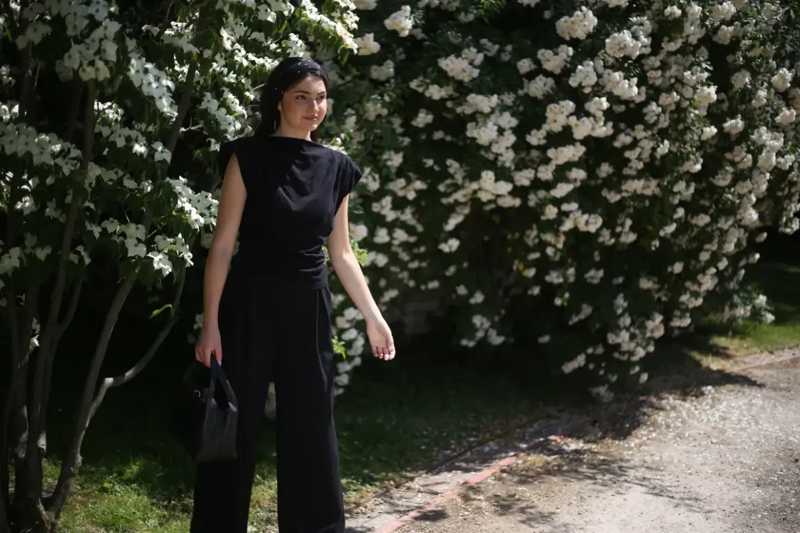 Kobieta w czarnym, eleganckim stroju, jak prac czarne pranie, stoi na tle kwitnących krzewów.