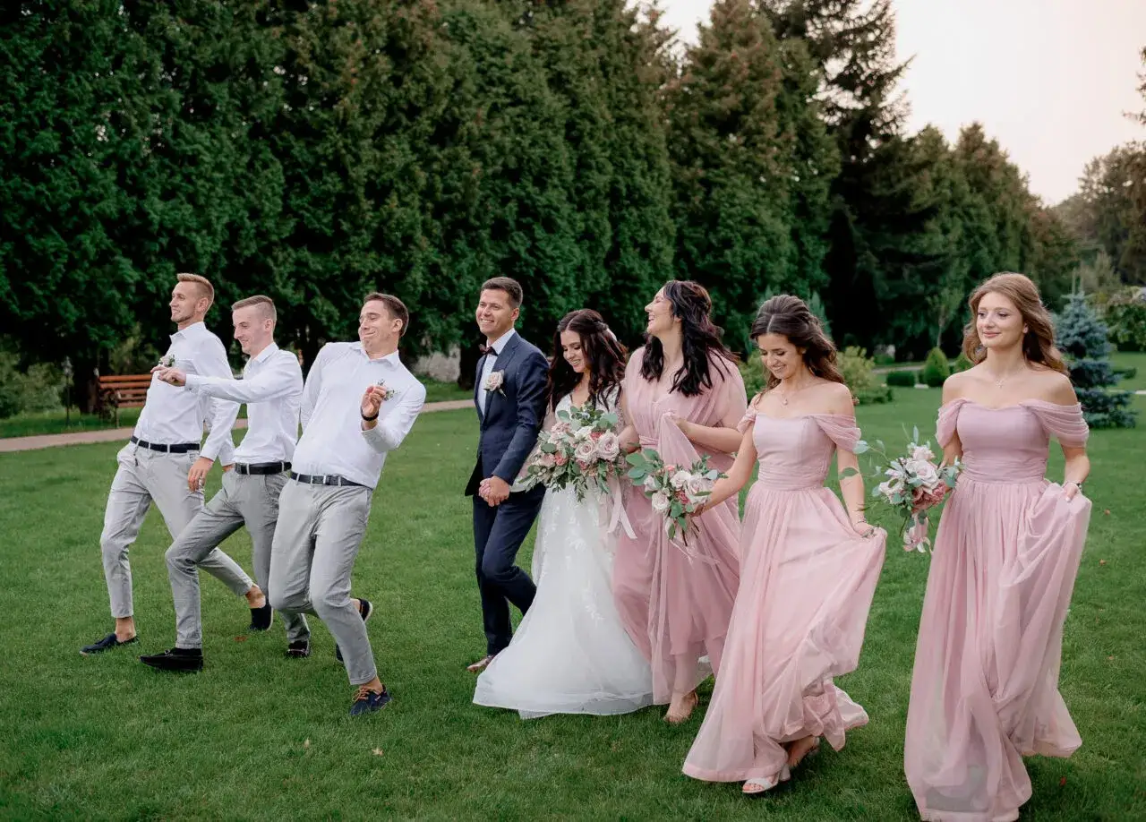 Uśmiechnięta para młoda i drużyna weselna w różowych sukniach i eleganckich stylizacjach na wesele spacerują po trawie.
