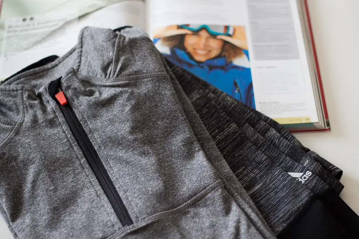Szara bluza sportowa z zamkiem i logo Adidas leży obok otwartej książki. Czy poliamid to dobry materiał na odzież sportową?