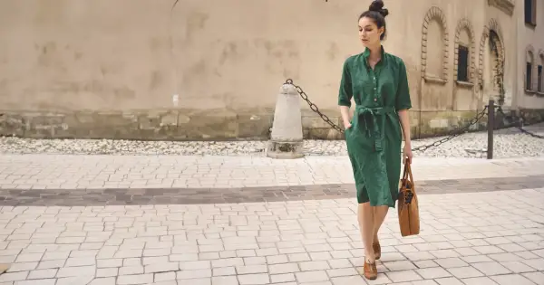 Elegancka kobieta w zielonej lnianej sukience prezentuje letnie stylizacje do biura, idąc ulicą z brązową torbą.