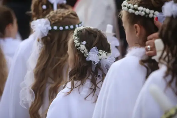 Dziewczynki w białych strojach z pięknymi fryzurami komunijnymi z wiankiem z kwiatów i wstążek.