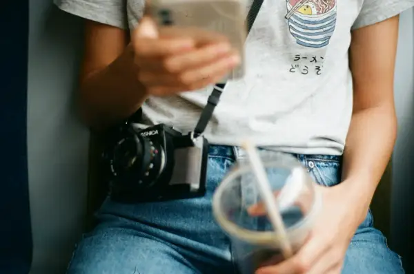 Osoba w jeansach z wysokim stanem (mid waist) trzyma telefon i napój. Na szyi wisi aparat.