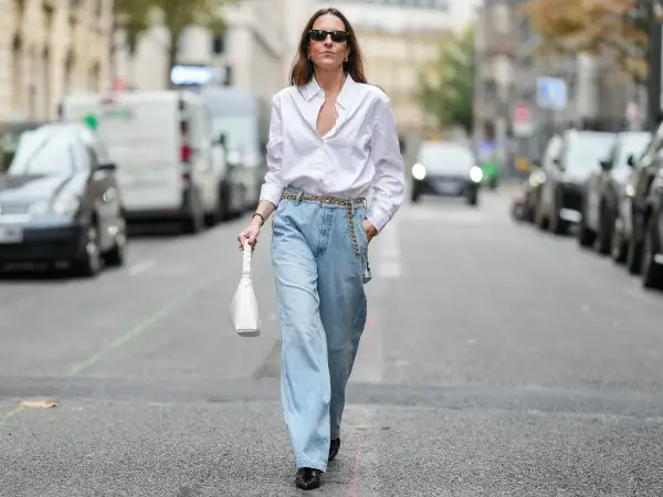 Kobieta w luźnych jeansach i białej koszuli, z białą torebką. To jest właśnie ten styl, który można nazwać "mom fit".